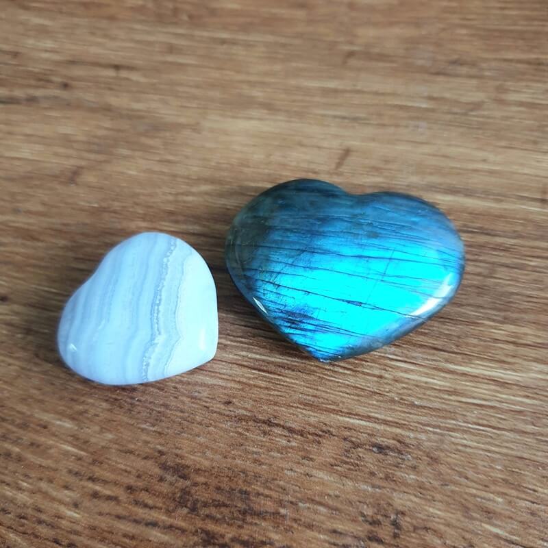 Zwei herzförmige Edelsteine, ein blau schimmernder Labradorit und ein hellblauer Chalcedon, liegen auf einer Holzoberfläche.