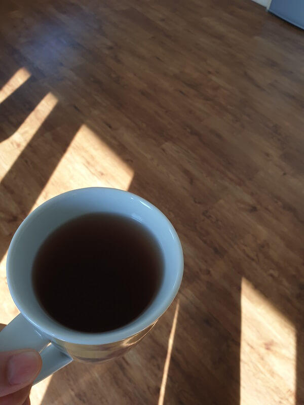 Blick von oben auf eine weiße Kaffeetasse in einer Hand, während helles Sonnenlicht lange Schatten auf einen Holzboden wirft.