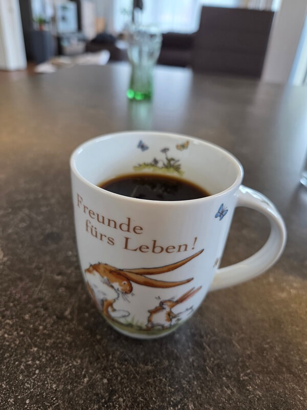 Eine Kaffeetasse aus Porzellan auf einem dunklen Küchentisch. Auf der Tasse steht „Freunde fürs Leben!“ mit einer Illustration aus „Weißt du eigentlich, wie lieb ich dich hab?“. Der Hintergrund ist unscharf.