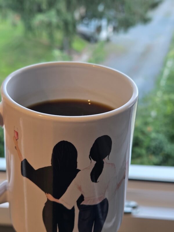 Nahaufnahme einer weißen Kaffeetasse am Fenster, gefüllt mit dunklem Kaffee. Das Motiv auf der Tasse zeigt zwei Frauen von hinten, die den Arm umeinander gelegt haben. Der Hintergrund durch das Fenster zeigt verschwommene Bäume im Garten.