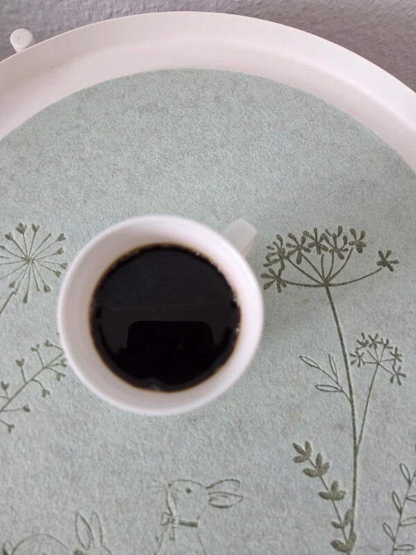 Draufsicht auf eine weiße Tasse mit schwarzem Kaffee auf einem blaugrauen Filz-Untersetzer mit aufgesticktem Hasen und Blumenmuster.