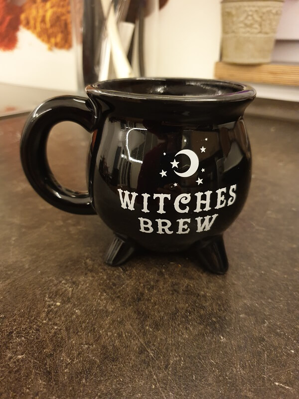 Eine schwarze Keramiktasse in Form eines Hexenkessels, bedruckt mit einem Mond, Sternen und dem Schriftzug „WITCHES BREW“.