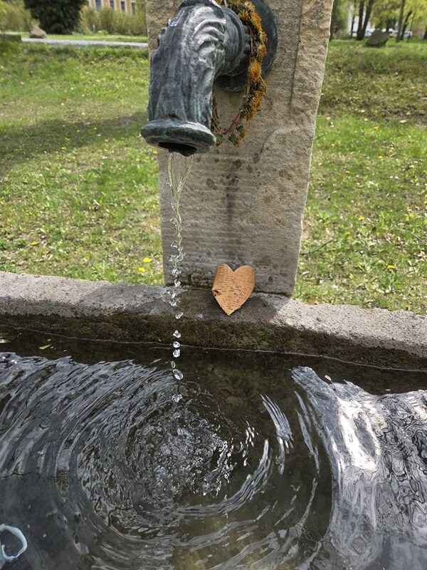 Ein Brunnen aus Stein, aus dem Wasser in ein Becken fließt. Am Fuß der Brunnensäule lehnt ein kleines Herz aus Rinde.