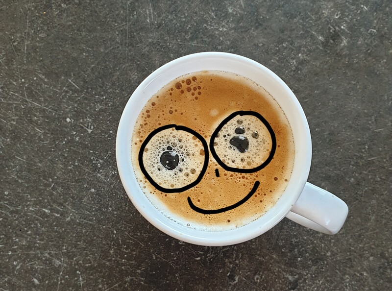Eine weiße Tasse Kaffee mit Crema, in die zwei dicke schwarze Kreise als Augen und ein lächelnder Mund gemalt sind.